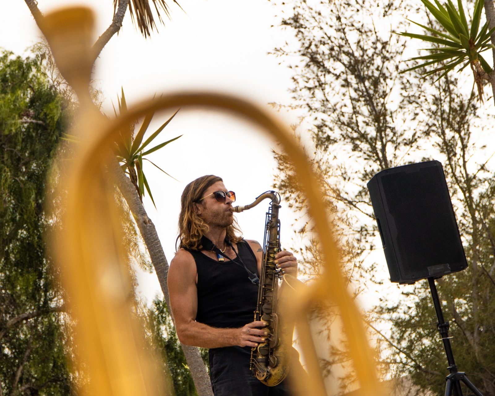 Luca Simonelli tocando el saxofón en un evento al aire libre, enmarcado a través de la campana de una trompeta